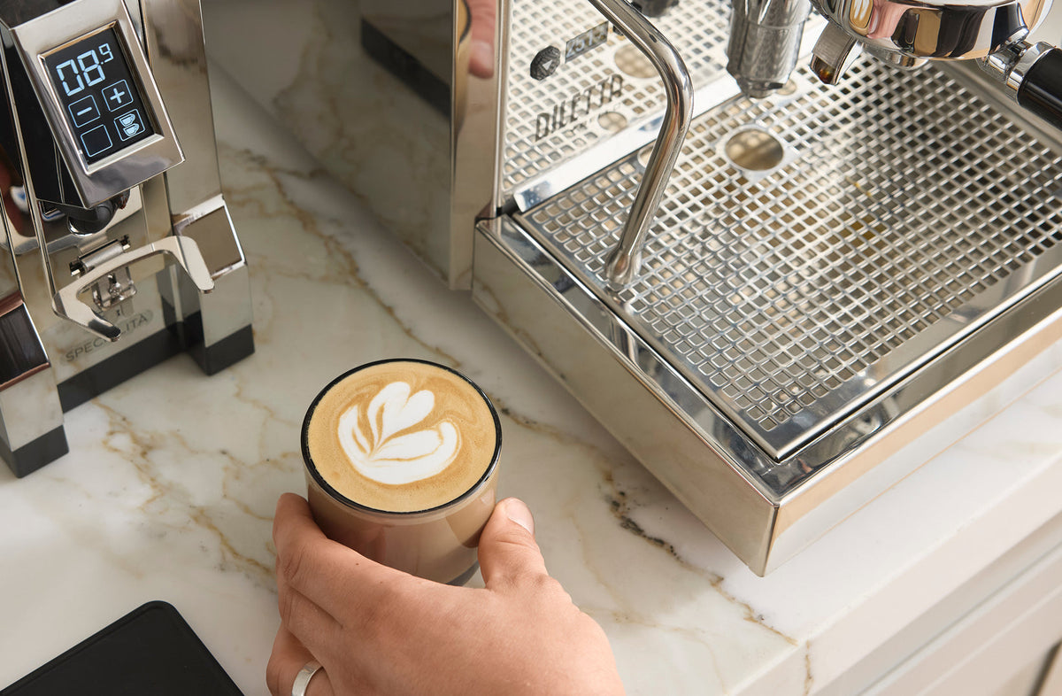 Hand holding a latte with latte art beside a Diletta espresso machine and Eureka grinder - Seattle Coffee Gear