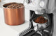 Fine espresso grind dosing into a portafilter from a burr grinder beside copper bean canister
