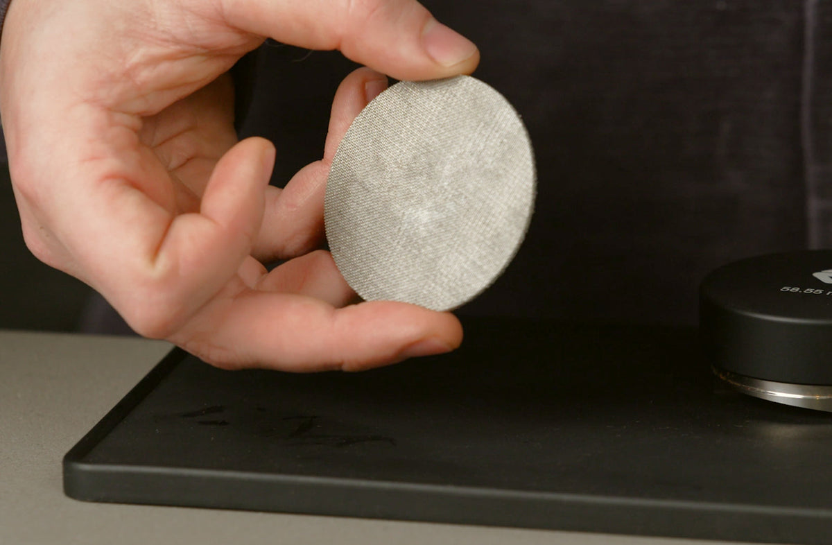 Barista holding a stainless steel espresso puck screen above a tamping mat
