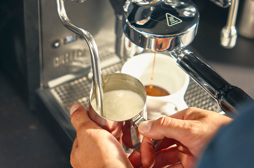 Hands holding a milk pitcher near a steaming espresso machine while a shot extracts into a white cup