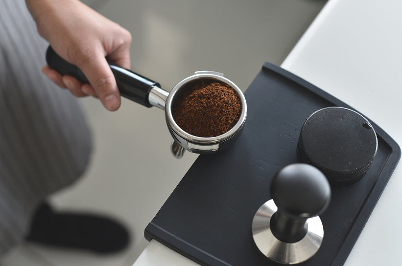 A portafilter basket filled with finely ground coffee next to a tamper on a tamping mat