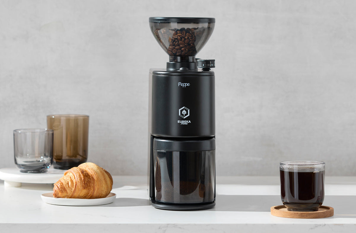 Matte black Eureka Peppe grinder on a kitchen counter beside brewed coffee and a croissant