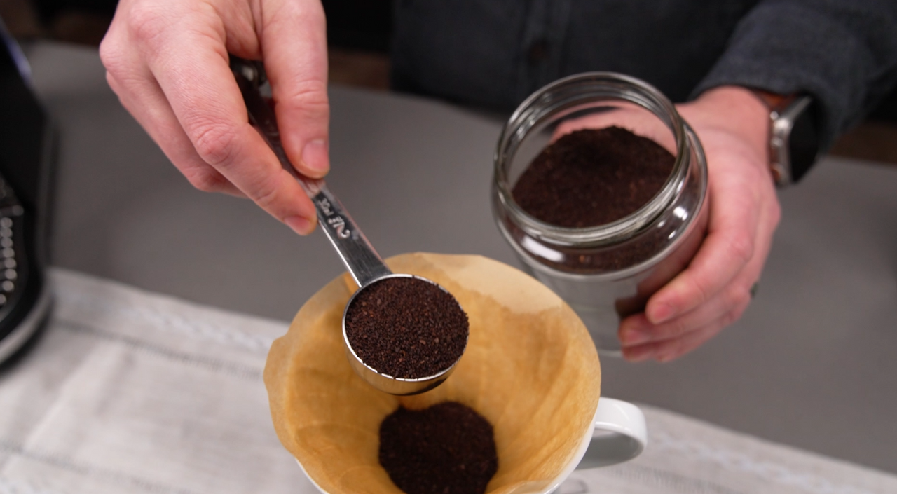 Scooping pre-ground coffee from a glass jar into a paper filter in a pour over dripper