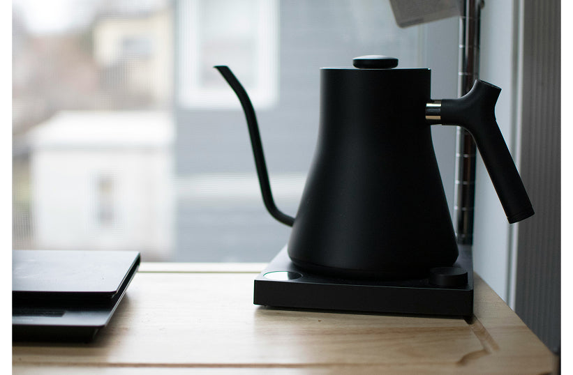 A matte black gooseneck electric kettle sitting on its base near a window